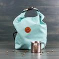 Load image into Gallery viewer, Seafoam green roll-top dog kibble travel bag with orange logo, styled with scattered kibble and a camping cup
