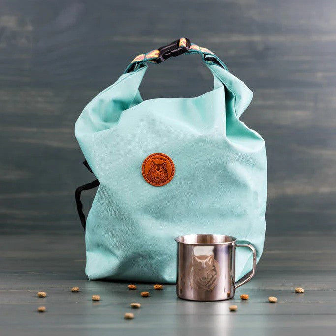 Seafoam green roll-top dog kibble travel bag with orange logo, styled with scattered kibble and a camping cup